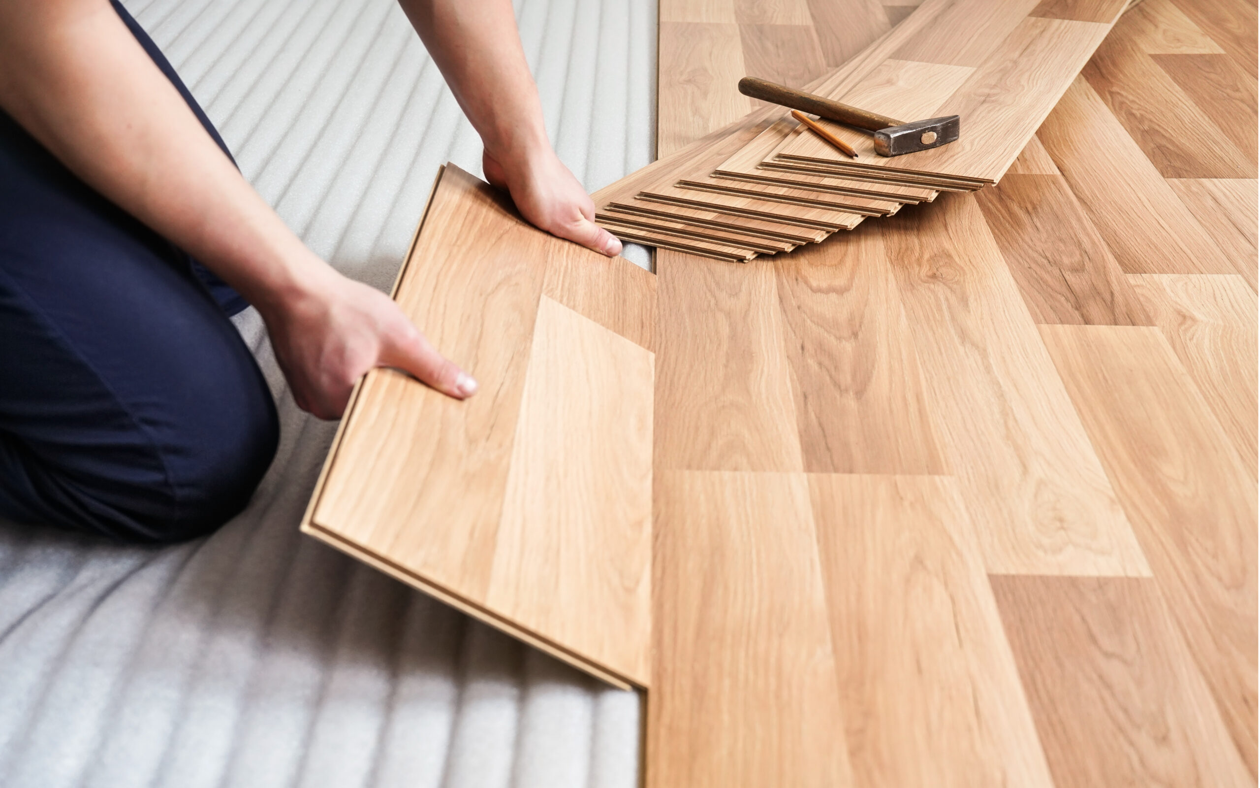 a person installing wood patterned laminated flooring with a hammer, a pencil and a stack of laminate floor tiles in the background